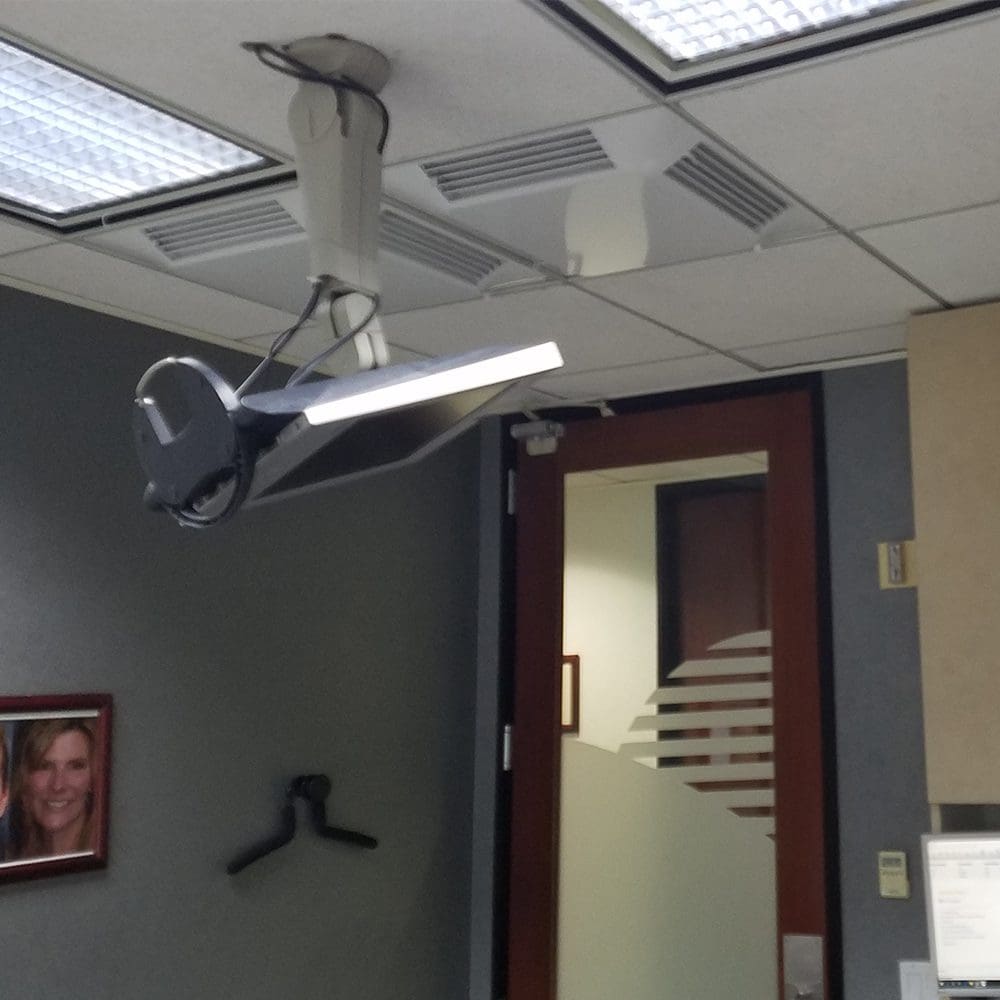 A ceiling-mounted Comfort First Filtered Diffuser is positioned above a dental chair in an office, with a framed photo on the wall and a door featuring a frosted glass design in the background.