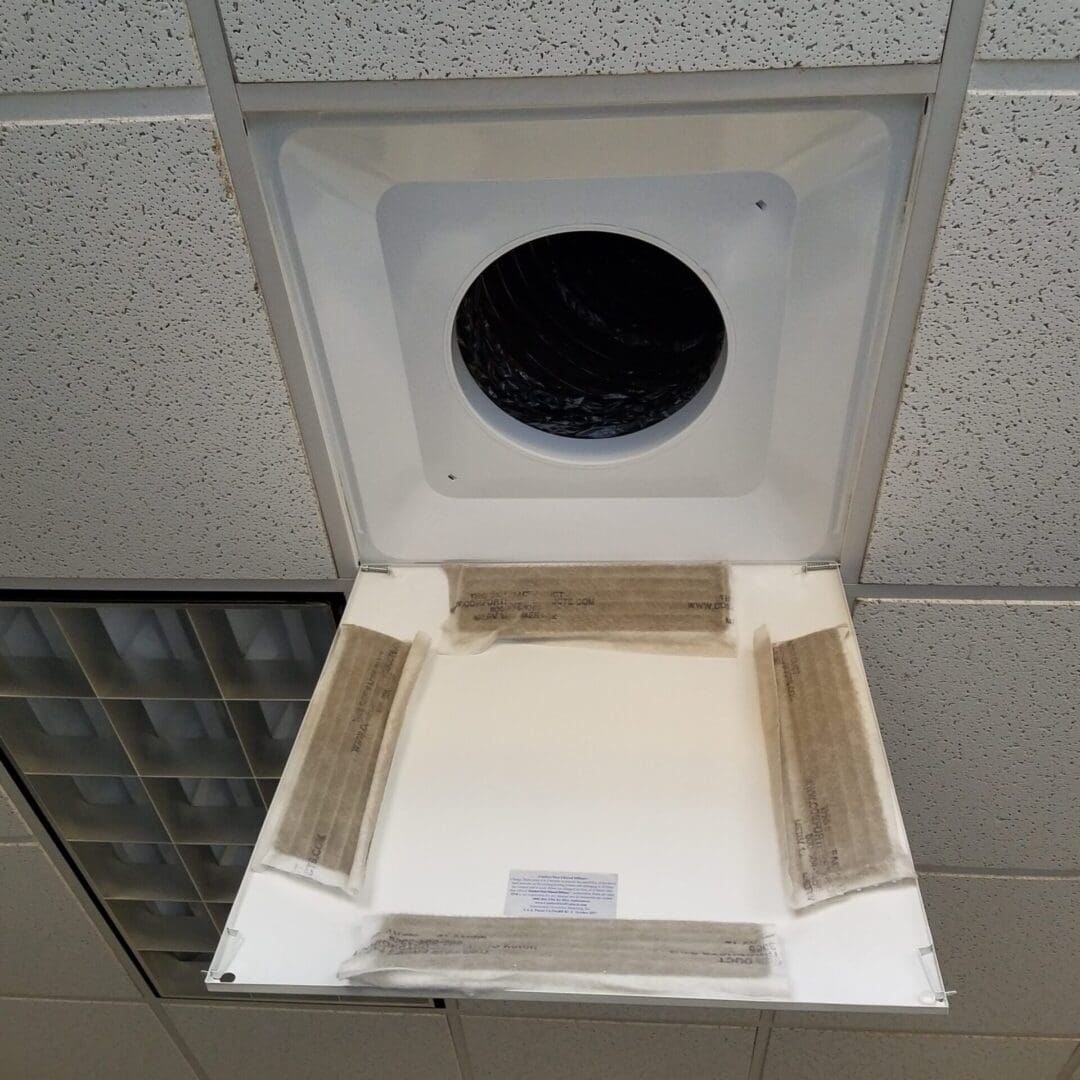 The Comfort First Filtered Diffuser, a commercial 4-way adjustable vent diverter, shows dust accumulation on its edges and is set in a drop ceiling with a hinged cover.