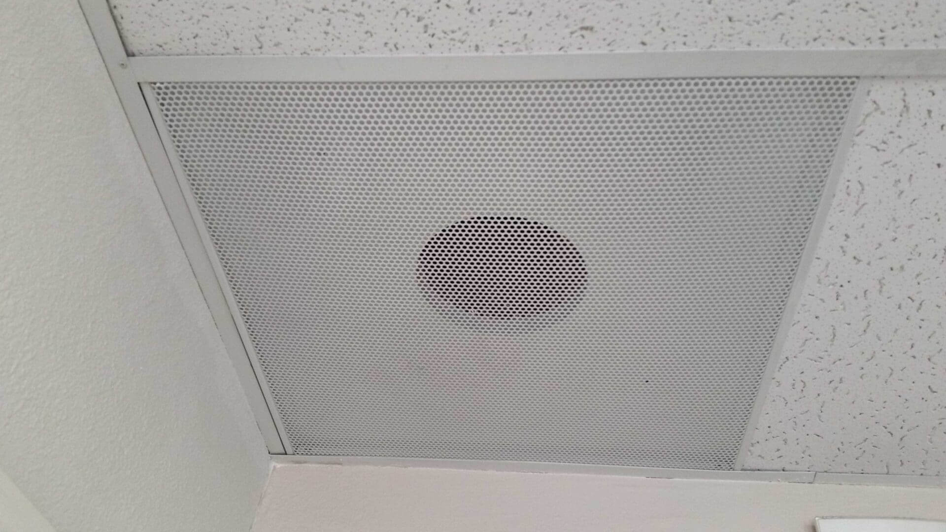 The Perforated Diffuser With Various Neck Sizes is a ceiling-mounted speaker embedded in an acoustic tile, surrounded by off-white textured ceiling tiles.