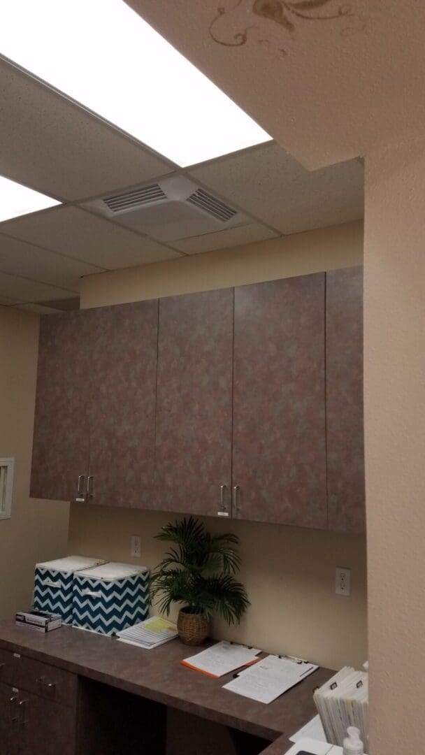 Office room featuring wooden cabinets, a countertop with papers, a potted plant, patterned boxes, ceiling lights, and the Comfort First Filtered Diffuser for optimal air distribution.